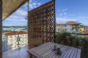 A balcony with a wooden table and chairs, a decorative metal screen, and a view of buildings and a body of water.