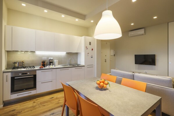 A modern kitchen with white cabinets, a built-in oven, and a dining area with a table and orange chairs.