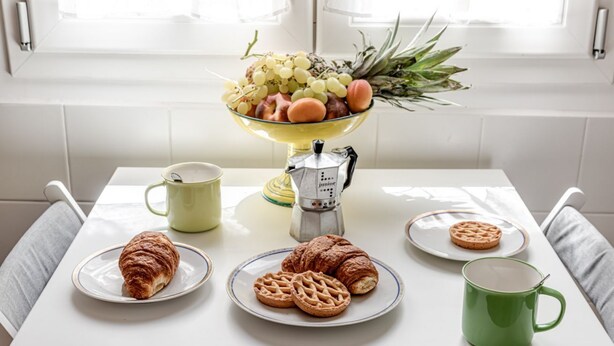 A breakfast setting with croissants, waffles, a green mug, a coffee pot, and a fruit bowl on a white table. - Serenity - Sweet Dreams in Florence (Firenze)