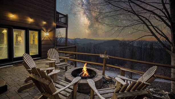 A wooden deck with Adirondack chairs and a fire pit, overlooking a mountainous landscape with a starry sky. - Private Sunset-view Cabin • Hot Tub • Sauna • Game Room • Dog-friendly (Blairsville)