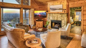 A cozy cabin interior with a stone fireplace, leather sofas, a television, and a scenic view through large windows. - Private Sunset-view Cabin • Hot Tub • Sauna • Game Room • Dog-friendly (Blairsville)