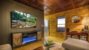 A cozy living room with a large flat-screen TV, a fireplace, wooden walls, and a view of a bridge and forest. - Private Sunset-view Cabin • Hot Tub • Sauna • Game Room • Dog-friendly (Blairsville)