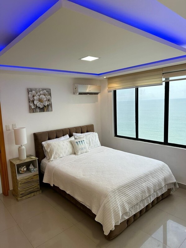 Ein Schlafzimmer mit Bett, Nachttisch, Klimaanlage, Fenster mit Blick und Deckenbeleuchtung in Blau.