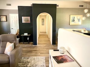 A modern living room with a sofa, a coffee table, and a bookshelf. There is a door leading to another room, and a hallway with a painting on the wall.