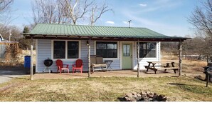 Una piccola casetta con portico coperto, due poltrone Adirondack rosse, un barbecue e un tavolo da picnic.