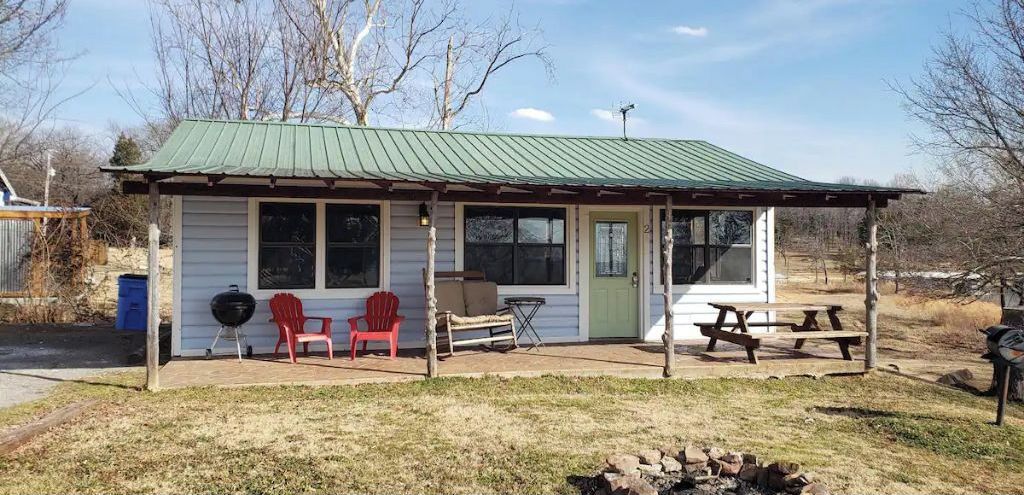 Een klein blokhut met een overdekte veranda, twee rode Adirondack-stoelen, een barbecue en een picknicktafel.