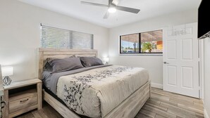 Un dormitorio con cama, mesita de noche, ventilador de techo, ventana con persianas y una puerta.