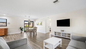 Una sala de estar moderna con un televisor blanco, sofás grises, una mesa de centro blanca y una zona de comedor con una mesa y sillas.