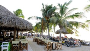 Ein Restaurant direkt am Strand mit strohgedeckten Gebäuden, Holztischen und -stühlen sowie Palmen im Hintergrund.