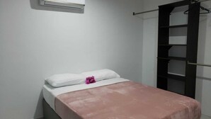 Ein kleines Zimmer mit einem Einzelbett, einer weißen Wand, einer braunen Bettdecke und einem weißen Kopfkissen.