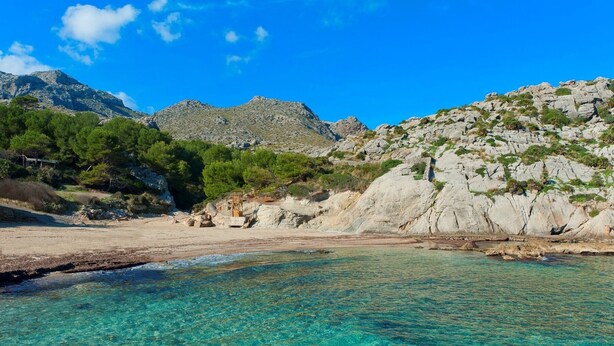 Una playa con agua turquesa cristalina, acantilados rocosos y vegetación escasa.