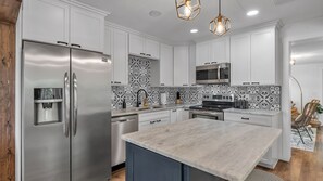 A modern kitchen with white cabinets, stainless steel appliances, a water dispenser, and decorative black and white tiles. - Modern Boho Retreat with Piano. Fenced and Gated in Central Florida! (Lakeland)