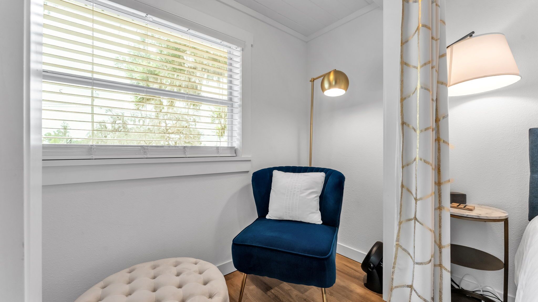A room with a window, a blue chair, a white pillow, a floor lamp, and a curtain with a gold pattern.