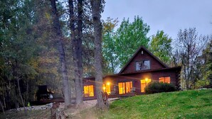 A cabin with a fire pit in the foreground and a well-lit house in the background.