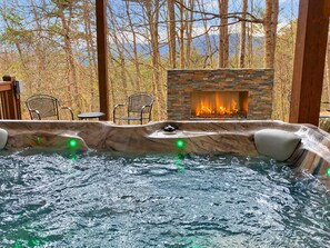 A hot tub with a stone fireplace, surrounded by chairs and a wooden deck. - Timber Ridge Falls | 7-Bedroom Asheville Vacation Rental with Hot Tub & Game Room (Fairview)