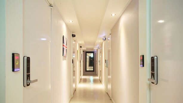 A modern hotel hallway with a long, straight layout, featuring doors with digital keypads and illuminated signs indicating room numbers. - Bucheon THE Play (Bucheon)