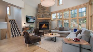 Una sala de estar amplia con chimenea de piedra, un televisor grande y una escalera que conduce a un nivel superior.