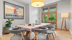 Comedor con una mesa dispuesta para seis personas, rodeada por seis sillas con estructura de madera y asientos de tela gris. Un cartel grande está en la pared sobre la mesa, y una lámpara sobre un pedestal a la derecha.