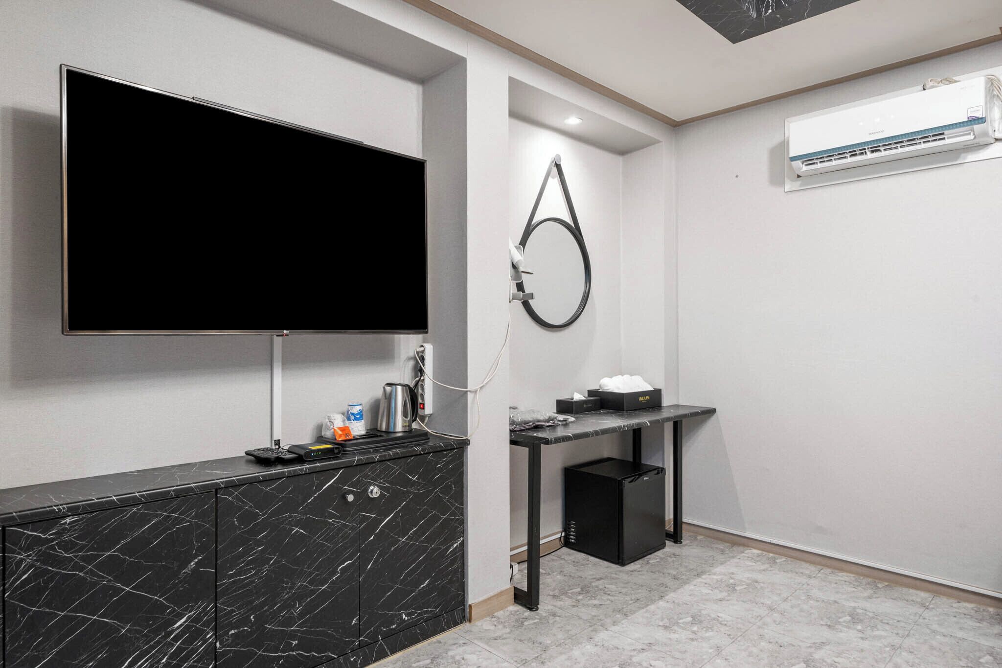 A modern kitchen with a black marble countertop, a flat-screen TV mounted above it, a small black refrigerator, and a wall-mounted air conditioner.