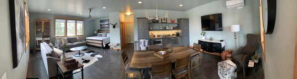 Uma sala de estar ampla, com um sofá grande, uma mesa de centro e uma mesa de jantar com cadeiras. Há uma cozinha com eletrodomésticos modernos e uma TV montada na parede.