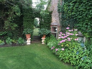 En trädgård med en stenlagd gång som leder fram till en stenvalv, omgiven av frodig grönska och blommande växter.