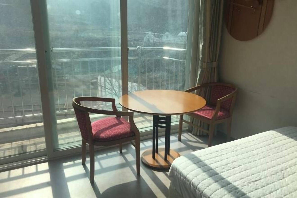 A room with a bed, a table, and two chairs. There is a large window with a view of buildings outside.