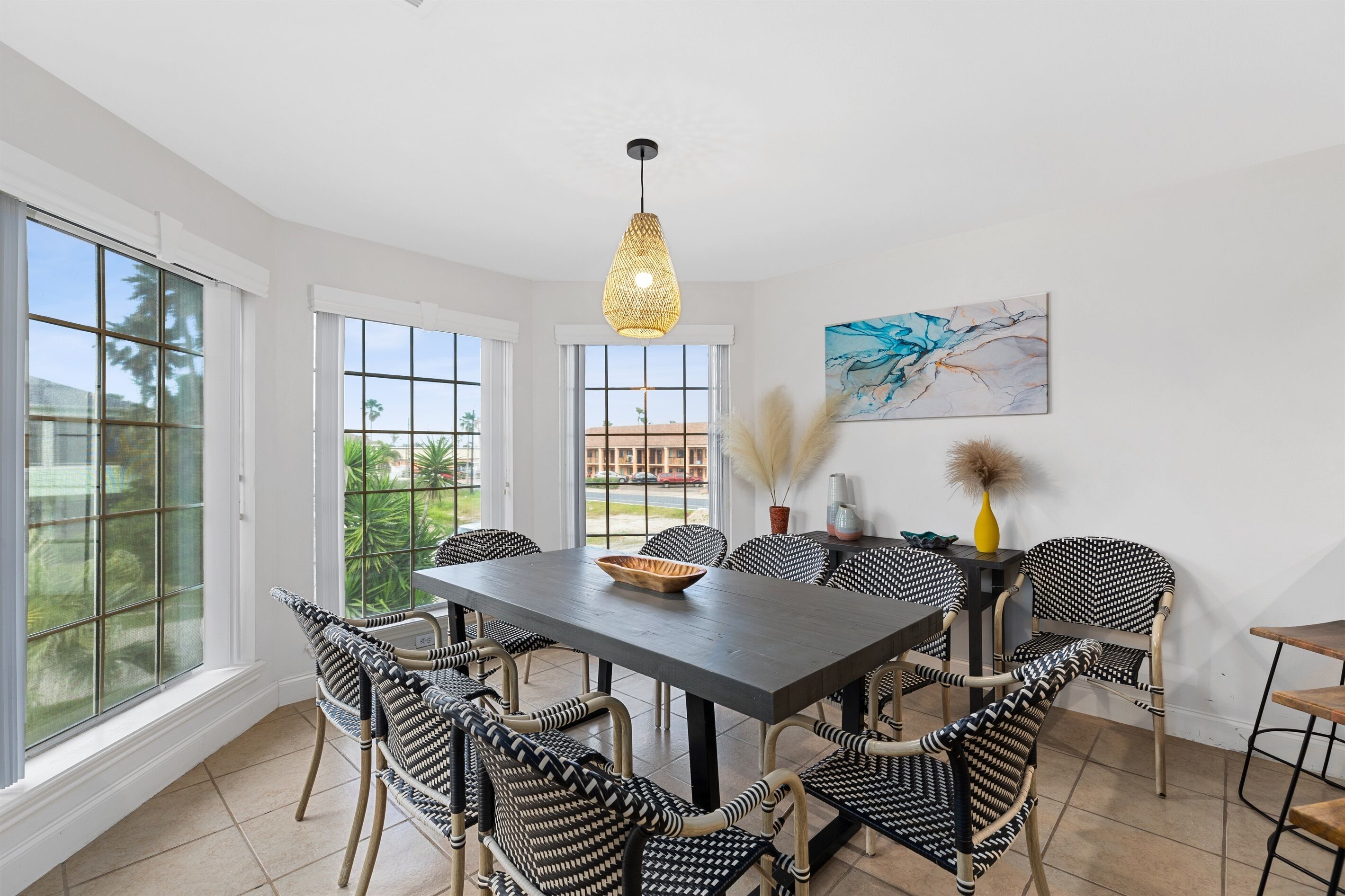 Un comedor con una mesa grande, sillas de diseño blanco y negro, una lámpara colgante tejida y vistas amplias a través de grandes ventanales.