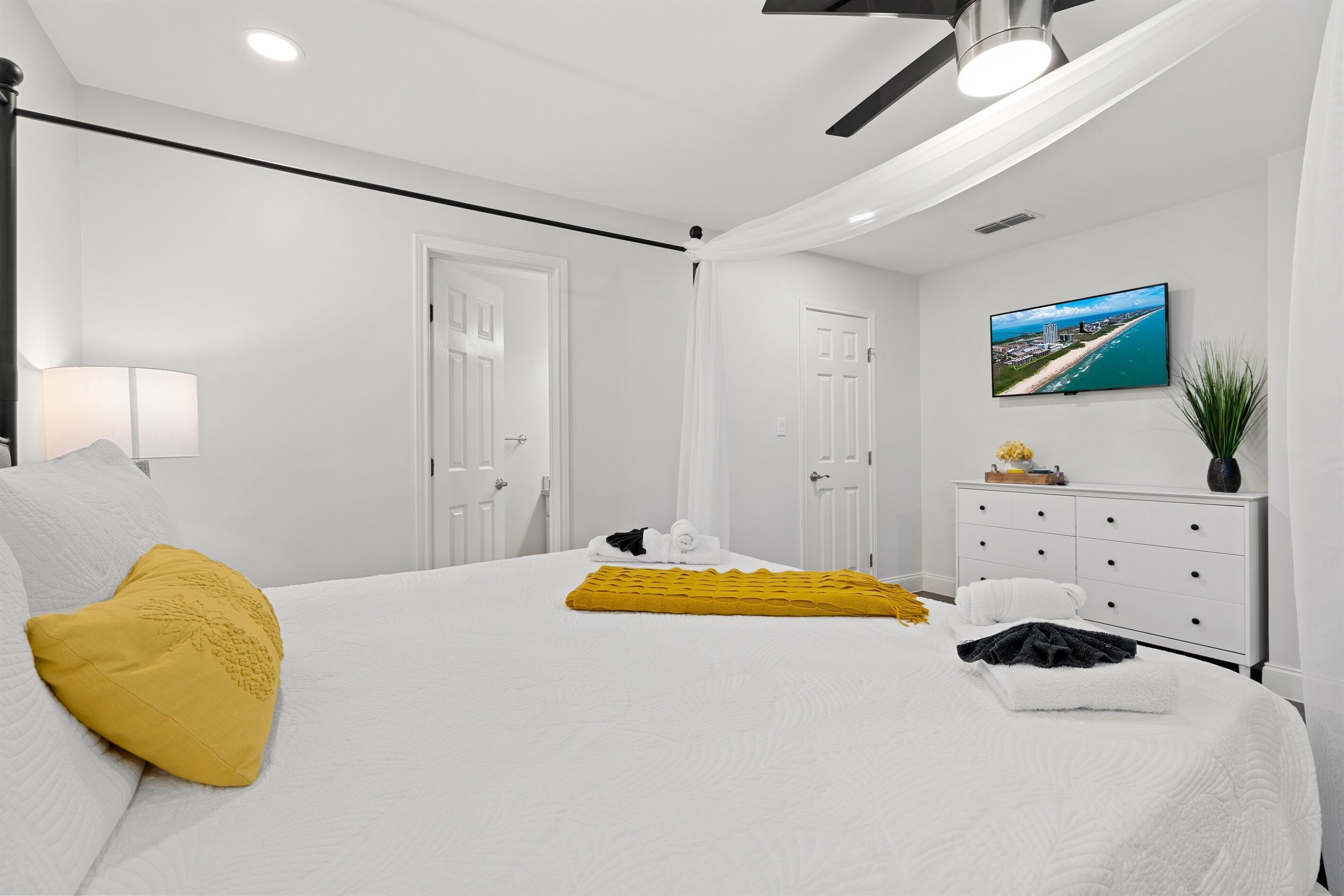 Habitación de hotel con cama, paredes blancas, ventilador de techo, televisor montado en la pared y ventana con vistas a la playa.