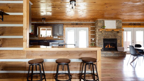 L’intérieur d’un chalet rustique, avec une cheminée en pierre, des tabourets de bar en bois et un coin cuisine doté d’une fenêtre donnant sur la terrasse.