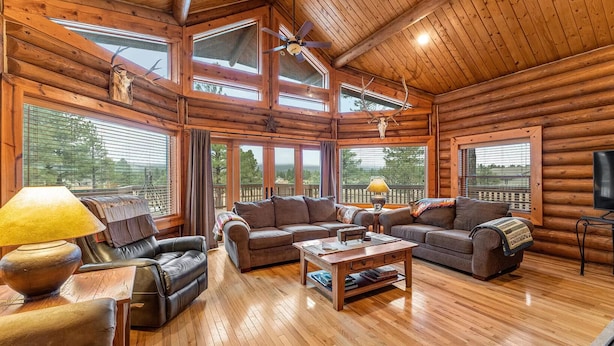 A log cabin interior with a wooden floor, leather furniture, a ceiling fan, and large windows offering a view of the outdoors. - Cozy Custom Cabin In The Pines-Williams AZ (Williams)