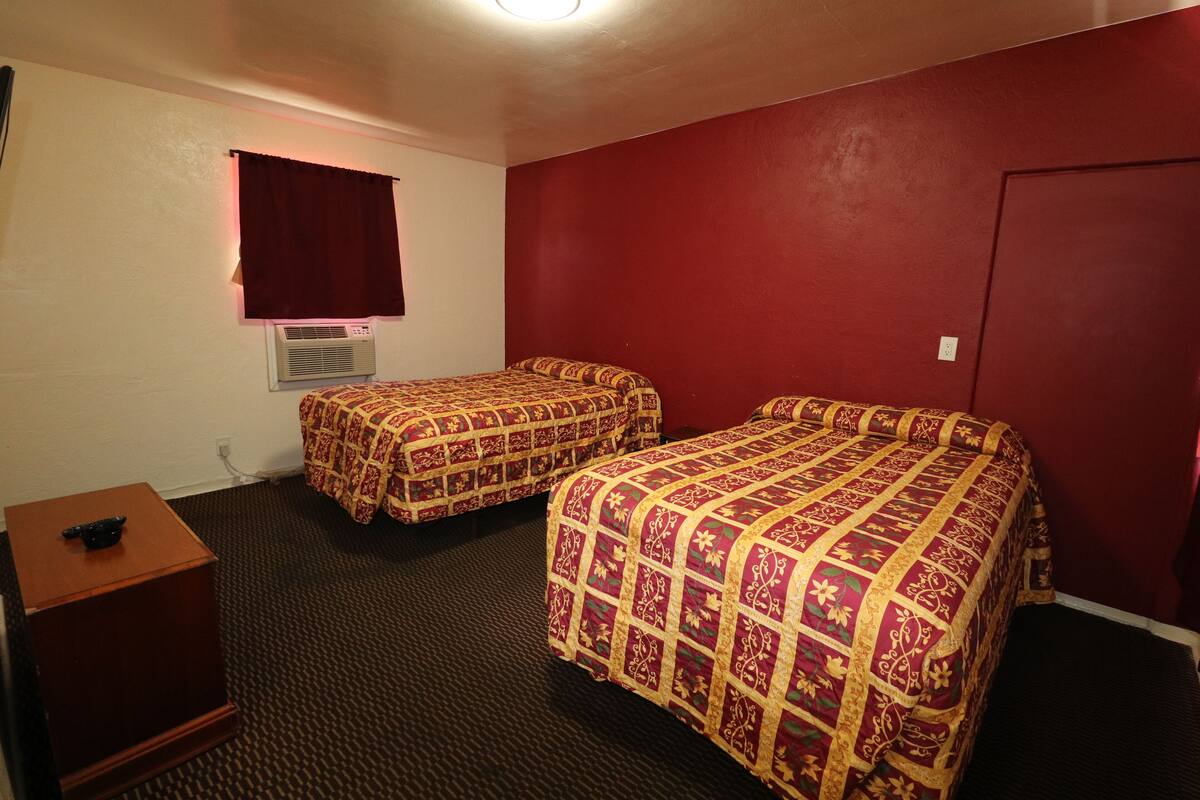 A hotel room with two beds, a wooden nightstand, a window with curtains, an air conditioning unit, and a door.