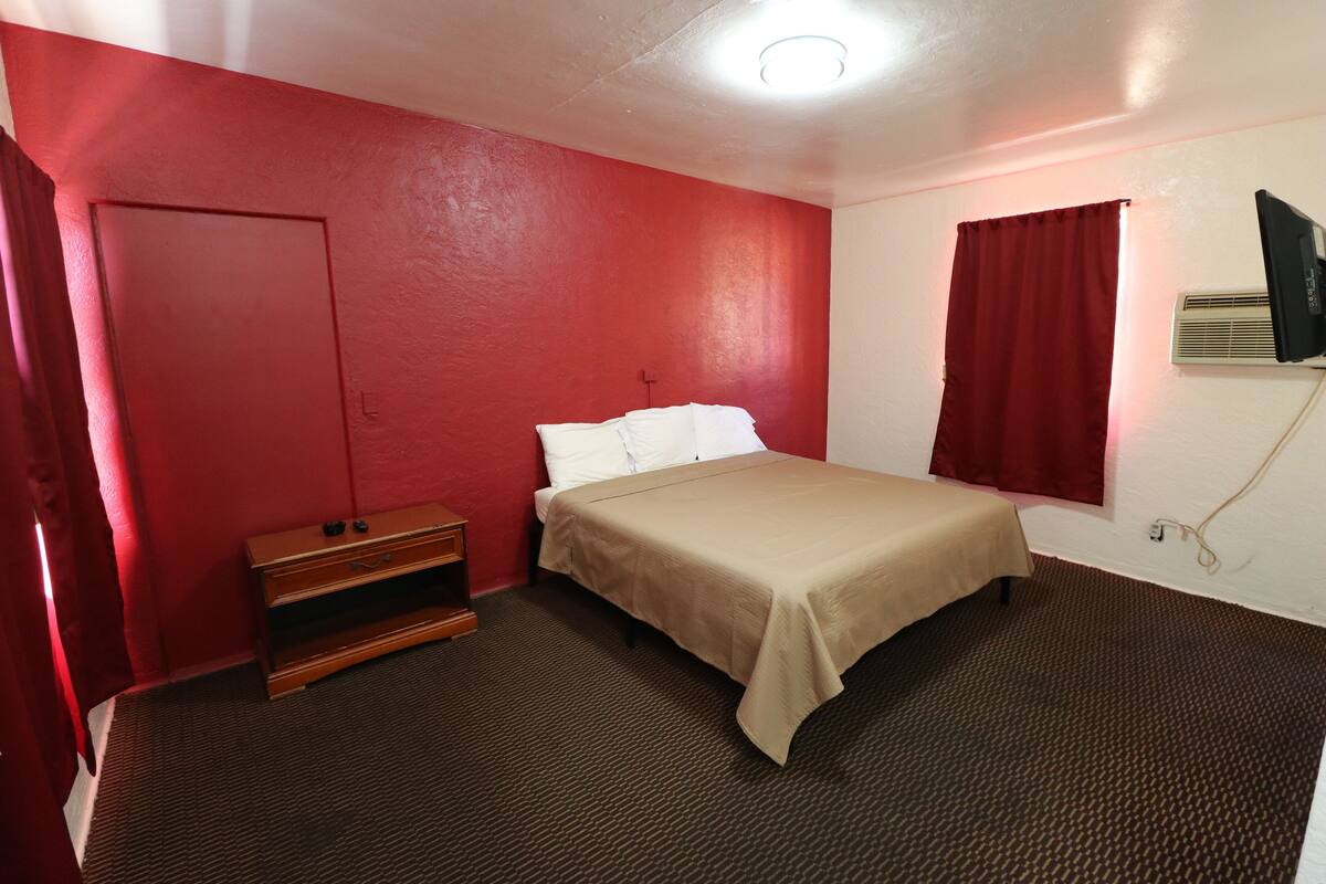 A room with a bed, a wooden dresser, a television mounted on the wall, and a window with red curtains.