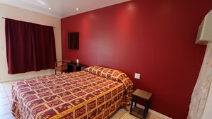 Ein Schlafzimmer mit roter Wand, einem Bett mit gemustertem Überwurf, einem Stuhl, einem kleinen Tisch und einem Fenster mit Vorhängen.