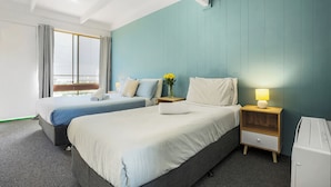 A hotel room with two beds, a nightstand with a lamp, a small table with a vase of flowers, and a view of the city through the window. - Coast Motel Eden (Eden)