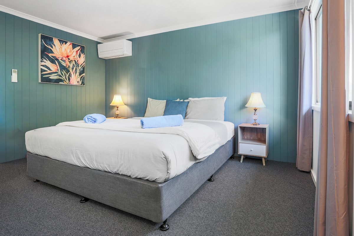 Ein ordentlich bezogenes Bett mit grauem Kopfteil, zwei Nachttische mit Lampen, ein Blumenbild an der Wand und ein Fenster mit Vorhängen.