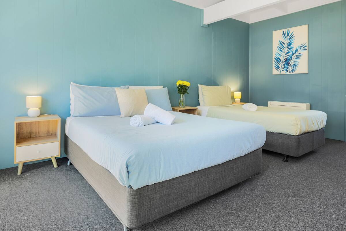 Ein Hotelzimmer mit zwei Betten, einem Nachttisch mit Lampe, einer Vase mit Blumen und einem gerahmten Bild an der Wand.