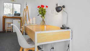A modern, well-lit room with a wooden desk, a white chair, a vase with red flowers, a black desk lamp, and a small stool. - Coast Motel Eden (Eden)
