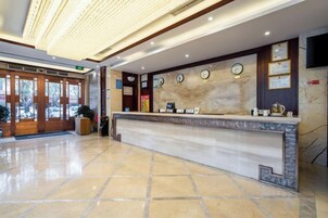 Zona de recepción de un hotel con mostrador de mármol, relojes y un televisor en la pared.