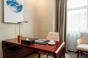 A hotel room with a wooden desk, a white chair, a cushioned armchair, a framed artwork, a bedside table, and a window with curtains. - Suiton Plus By Paxton Linzhi (Nyingchi)