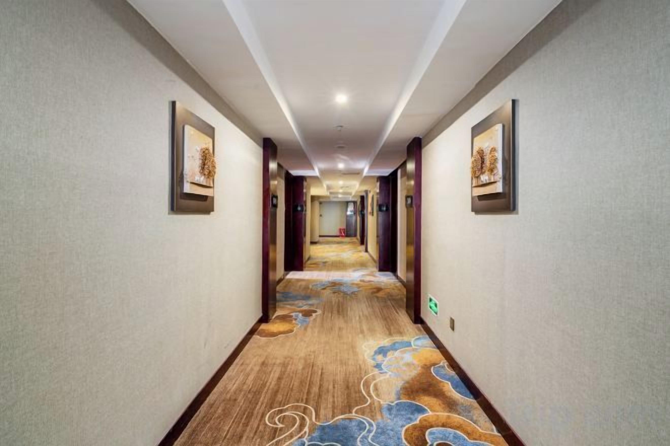 Un couloir d’hôtel avec un sol recouvert de moquette, des œuvres d’art encadrées accrochées aux murs et un luminaire au plafond.