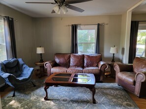 A living room with a brown leather sofa, a blue armchair, a wooden coffee table, and two lamps on side tables. - Cozy Fayetteville Home - King Bed - Sleeps 5 - Near Downtown (Fayetteville)