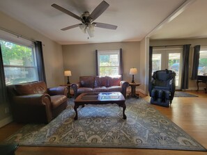 A living room with a ceiling fan, leather sofas, a coffee table, two lamps, and a view through the windows. - Cozy Fayetteville Home - King Bed - Sleeps 5 - Near Downtown (Fayetteville)