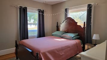 Een slaapkamer met een houten bed, twee kussens, een nachtkastje, een lamp, een plafondventilator en gordijnen.