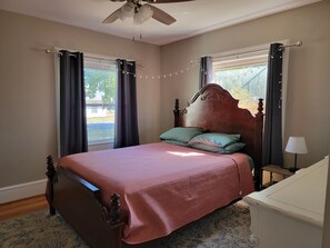 A bedroom with a wooden bed, two pillows, a nightstand, a lamp, a ceiling fan, and curtains. - Cozy Fayetteville Home - King Bed - Sleeps 5 - Near Downtown (Fayetteville)