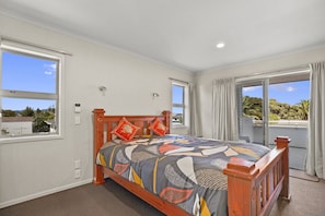 A bedroom with a wooden bed, large windows, and a view of the outdoors. - Endless Summer - Mount Maunganui Holiday Home (Tauranga)