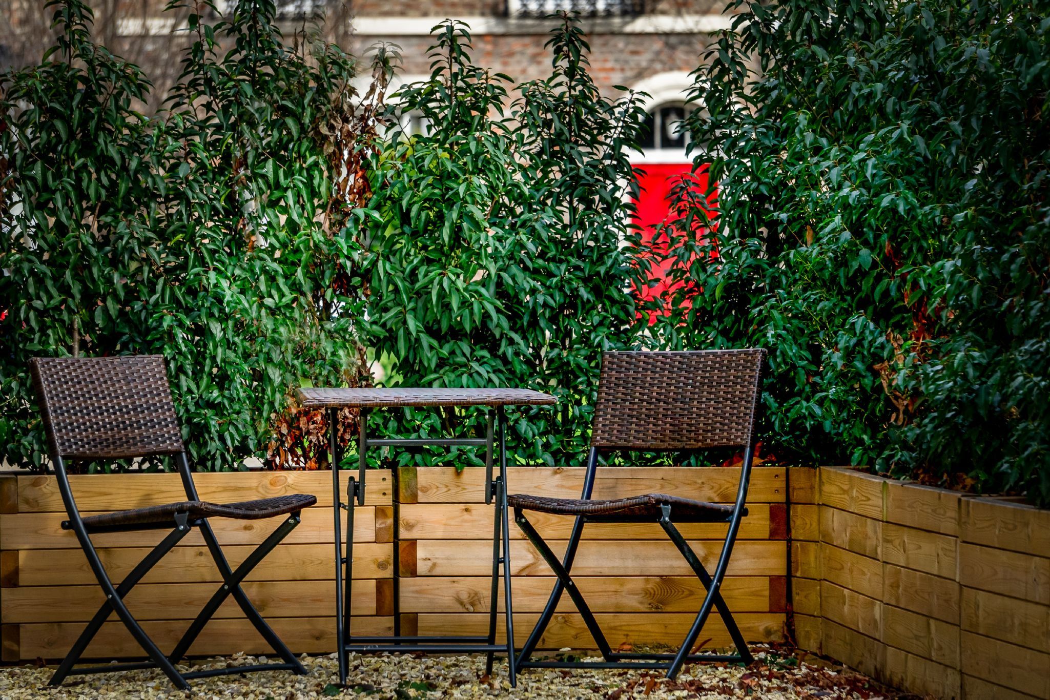 An outdoor seating area with two wicker chairs and a small table, surrounded by a wooden fence and dense foliage.