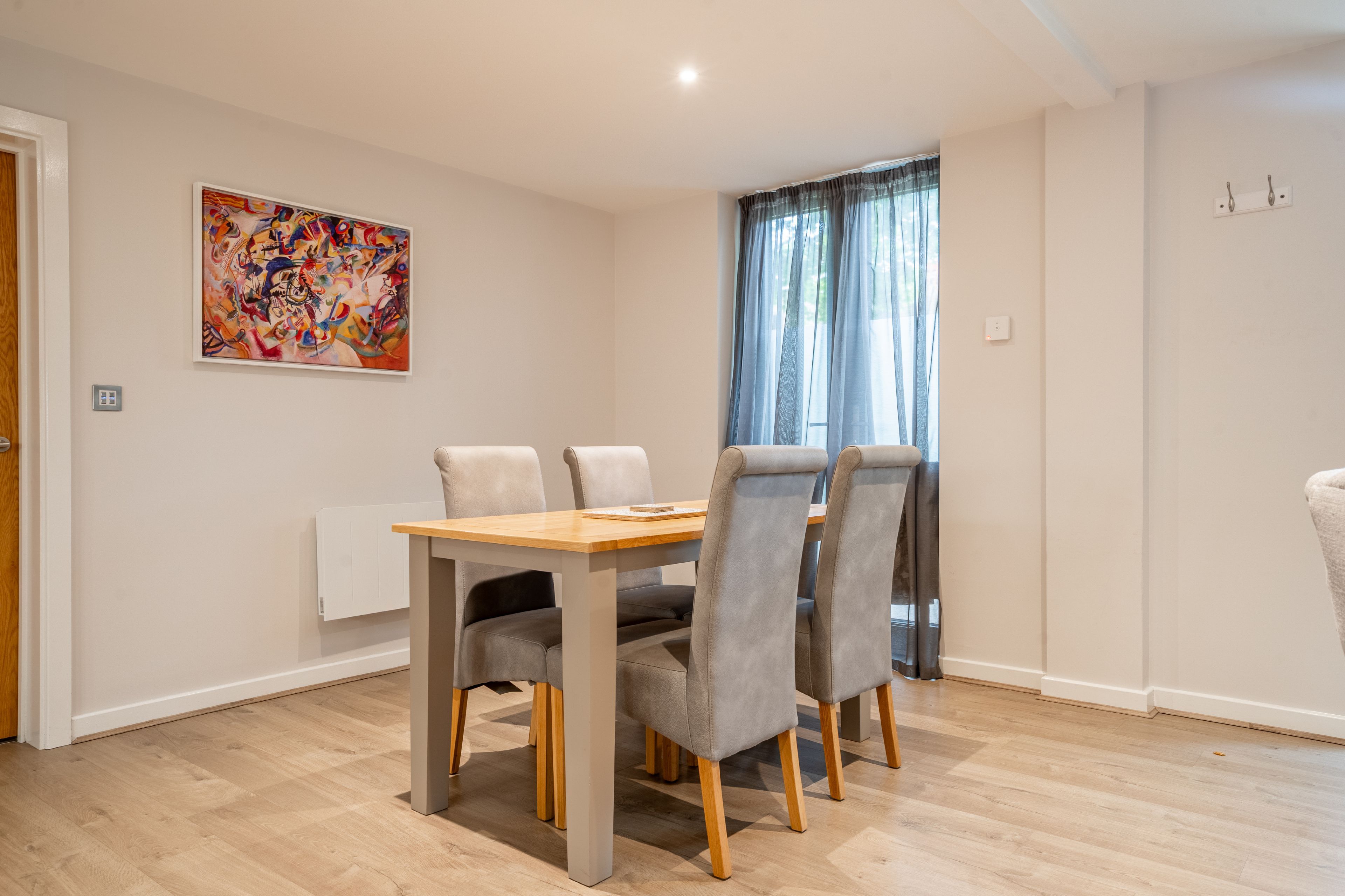 A dining area with a wooden table and four grey upholstered chairs, a radiator, a window with sheer curtains, and a colorful abstract painting on the wall.