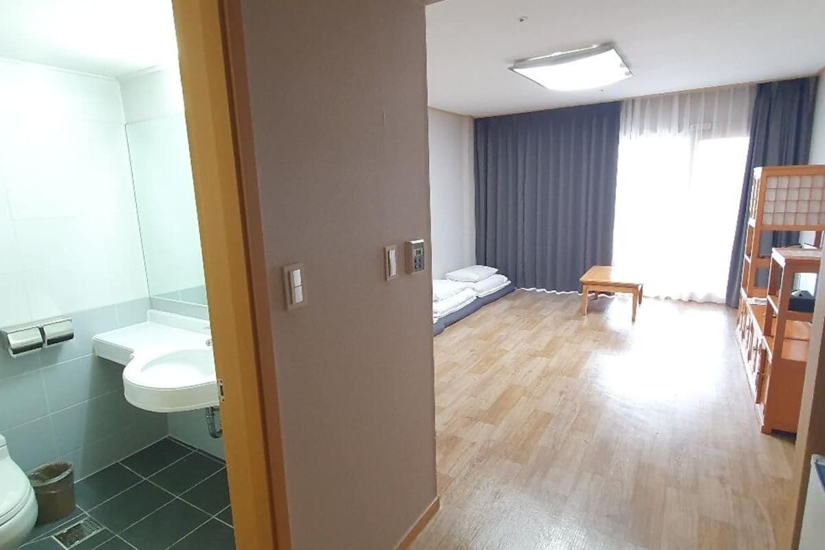 A hotel room with a wooden floor, a bed, a bedside table, a window with curtains, a bathroom with a toilet and sink, and a small wooden chair.