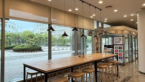 Een modern interieur met een lange houten tafel en barstoelen, grote ramen met uitzicht op de stad, en een automaat.