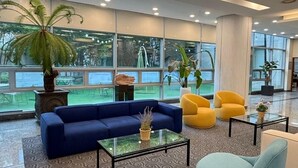 Een moderne lobby met een blauwe bank, gele stoelen, een glazen salontafel en grote ramen met uitzicht op het omliggende landschap.